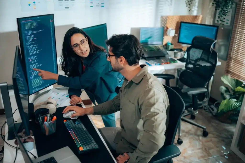 Two coworkers review code on a tall monitor, one pointing as they discuss software with a second screen and keyboard nearby.